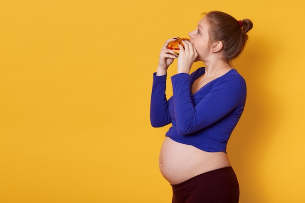 Side view of pregnant lady eats fast food, wearing blue shirt and leggins, with bunch on head