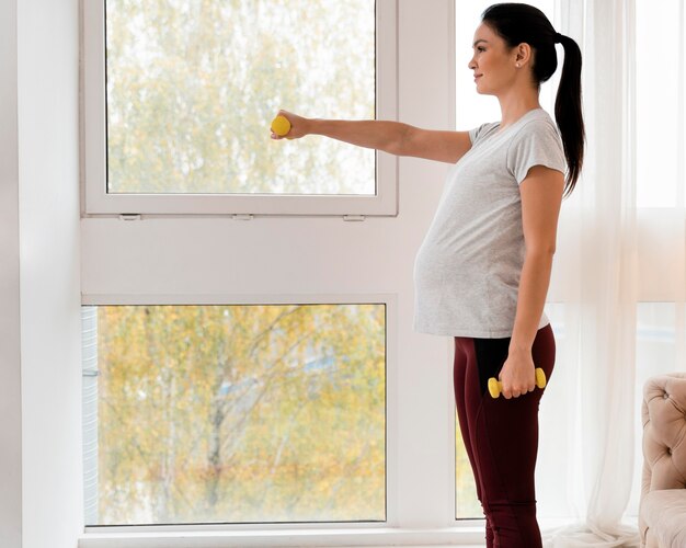 Вид сбоку беременная женщина, тренирующаяся с весами с копией пространства
