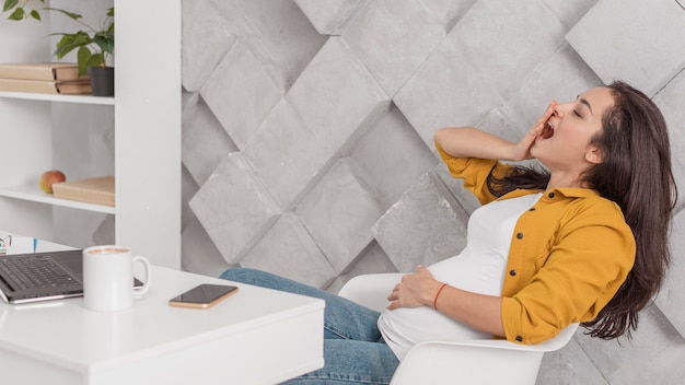 Side view of pregnant woman yawning at home with laptop and smartphone