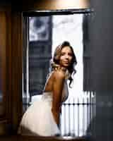 Free photo side view of pretty and pensive bride in ultrasexy wedding dress with thin straps which bending body forward looking away while posing on background of wooden wall and blur room