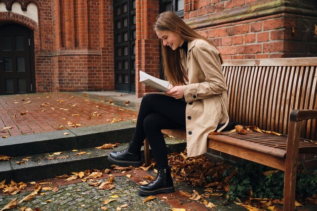 Side view of pretty stylih girl joyfully reading newspaper on bench on city street