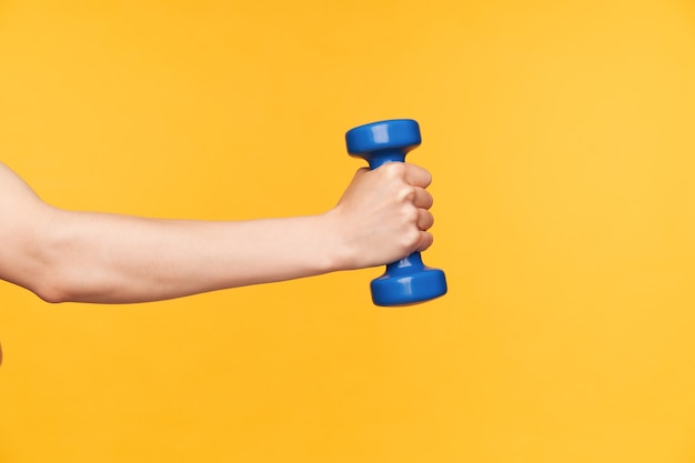 Side view of raised female hand with blue dumbbell exercising biceps while being isolated over yellow background. body care and fitness concept