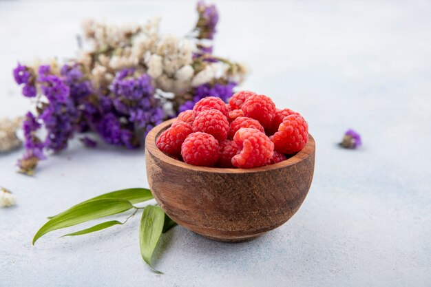 Side view of raspberries in bowl with flowers and leaves on white
