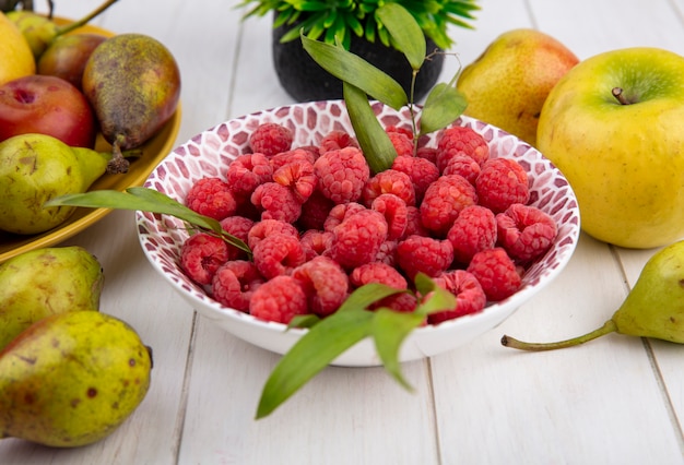 Side view of raspberries and leaves in bowl with peach and apple around on white