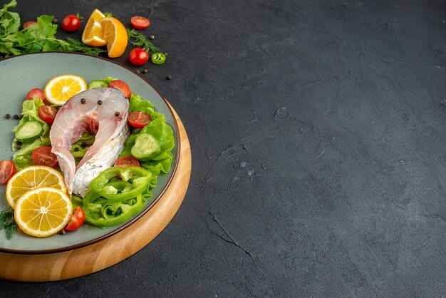 Side view of raw fish and fresh chopped vegetables lemon slices spices on a gray plate on a round board on black distressed surface
