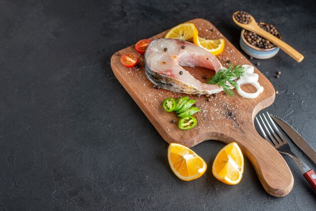 Side view of raw fish and fresh chopped vegetables lemon slices spices on a wooden board cutlery set on black distressed surface