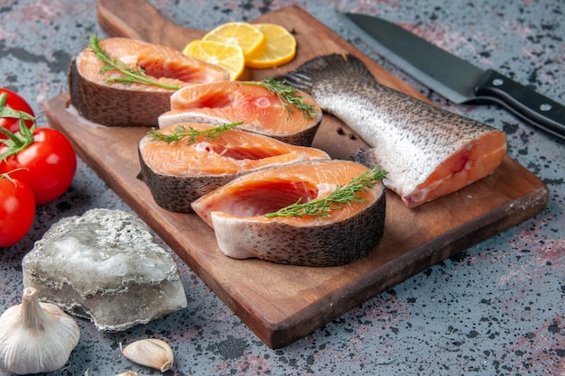 Side view of raw fishes lemon slices greens pepper on wooden cutting board and vegetables knife on blue black colors table