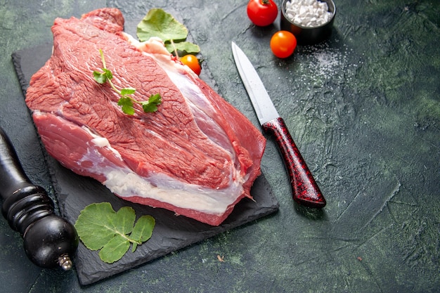 Side view of raw fresh red meat and greens on cutting board knife tomatoes wooden hammer on green black mix colors background