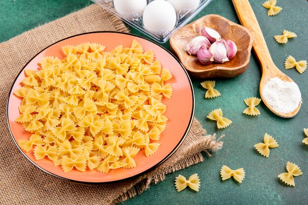 Side view of raw pasta on a plate with garlic eggs and a spoon with flour on a green surface