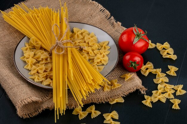 Side view of raw pasta with raw spaghetti and tomatoes on a beige napkin on a black surface