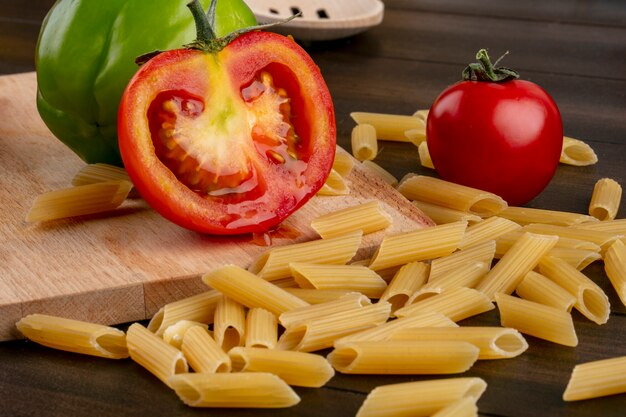 Side view of raw pasta with tomatoes and bell pepper on a cutting board on a wooden surface