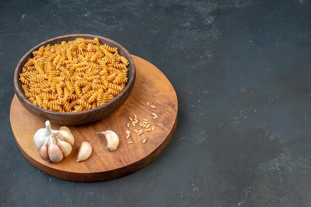 Side view of raw pastas in a brown bowl garlics rice on wooden board on the right side on black background