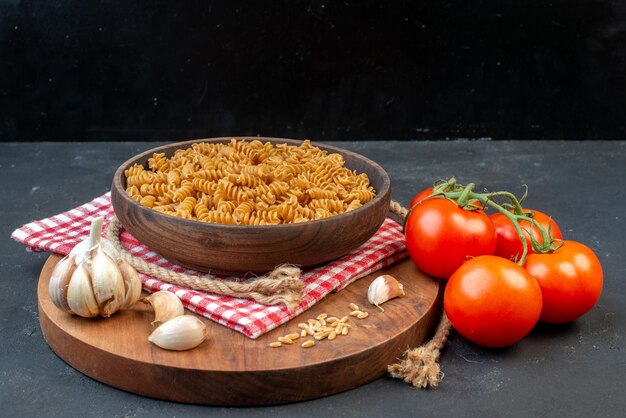 Side view of raw pastas in a brown bowl on red stripped towel garlics rice on round wooden board tomatoes with stem rope