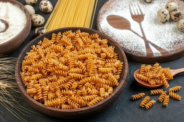 Side view of raw pastas in a brown bowl and spikes fork knive drawn with flour on cutting board flour on black background