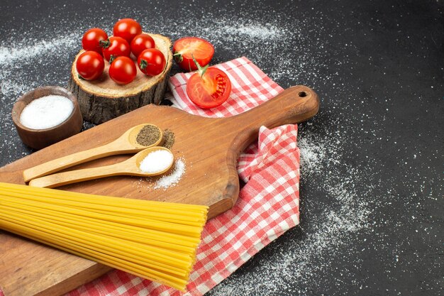 Вид сбоку на сырые макароны, соль, перец в ложках на разделочной доске на красном полосатом полотенце, целые нарезанные помидоры с правой стороны на черно-белом фоне