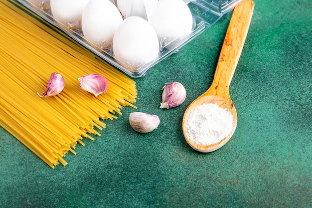 Side view of raw spaghetti with garlic and eggs with a wooden spoon with flour on a green surface