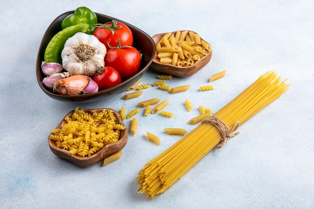 Vista laterale di spaghetti crudi con pasta cruda in ciotole con aglio e pomodori in una ciotola su una superficie grigia