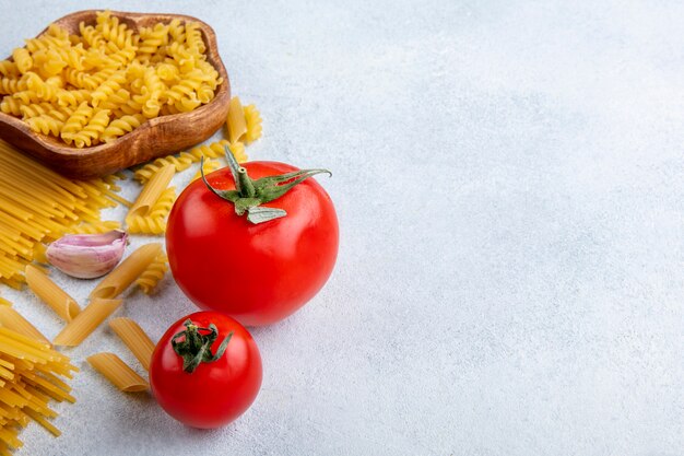 Side view of raw spaghetti with raw pasta in bowls with garlic and tomatoes on a gray surface