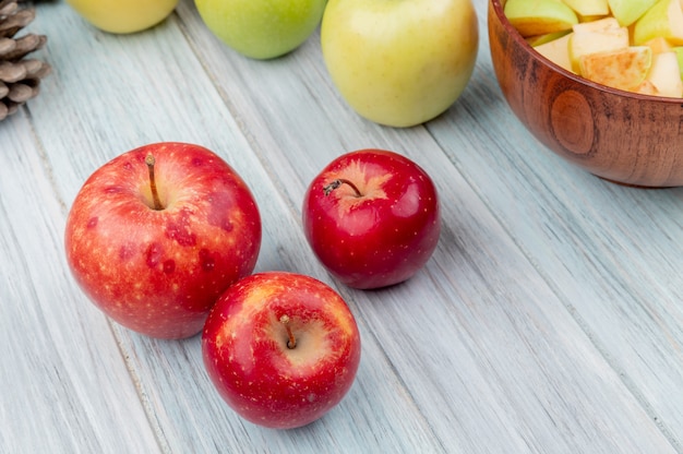 Вид сбоку красных яблок с желтыми и зелеными и миска ломтиков яблока на деревянном фоне