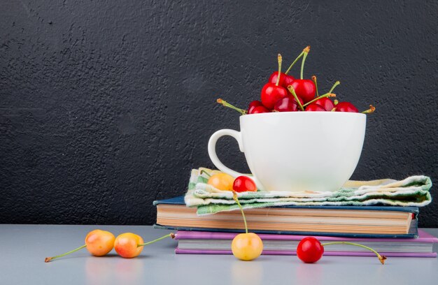 Side view of red cherries in cup on cloth and books with yellow cherries on blue surface and black background