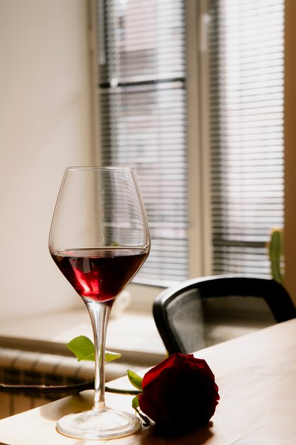 Вид сбоку розы красного цвета лежал возле бокала красного вина на деревянном столе на фоне окна