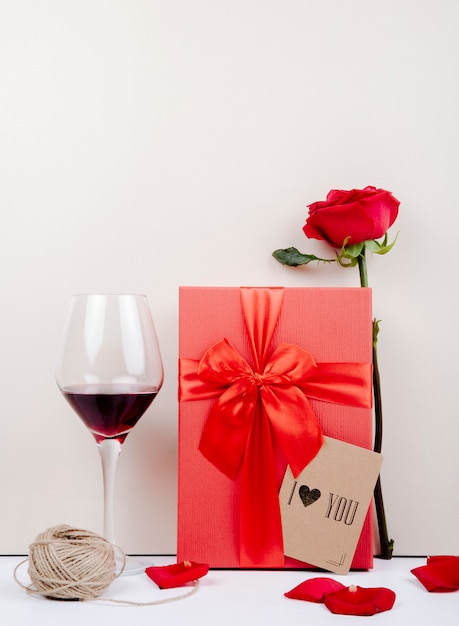 Side view of a red gift box with a bow and red rose with small postcard and a glass of wine a ball of rope on white background