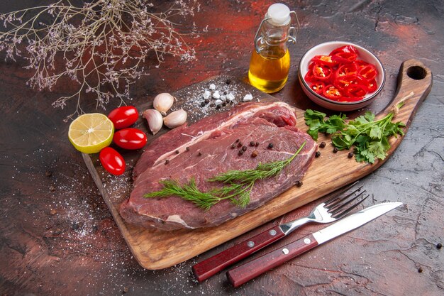 Side view of red meat on wooden cutting board and garlic green lemon chopped pepper tomato oil bottle on dark background