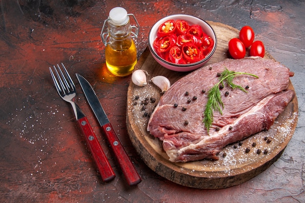 Side view of red meat on wooden tray and garlic green and chopped pepper fallen oil bottle fork and knife on dark background
