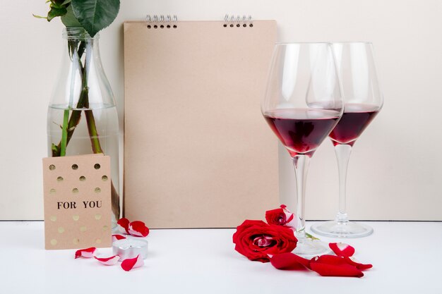 Side view of red roses in a glass bottle standing near a sketchbook and two glasses of red wine on white background
