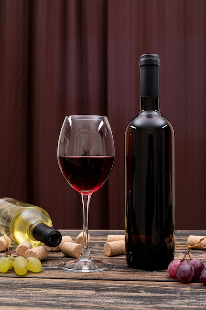 Вид сбоку красное вино в бутылке, стакан и виноград на темном столе и вертикали