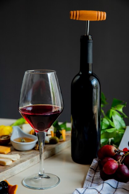 Side view of red wine bottle with corkscrew and different kinds of cheese olive walnut grape on white surface and black background