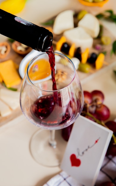 Side view of red wine pouring into glass and different kinds of cheese olive walnut grape and love card on white background