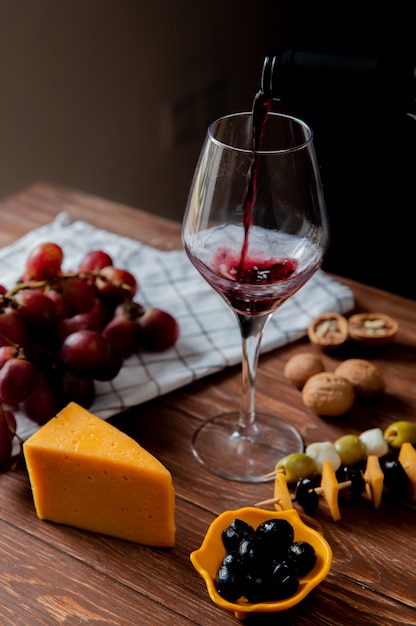 Вид сбоку красного вина, льющегося в стакан с чеддером и сыром пармезан, оливковое, виноград грецкого ореха на деревянной поверхности и черном фоне