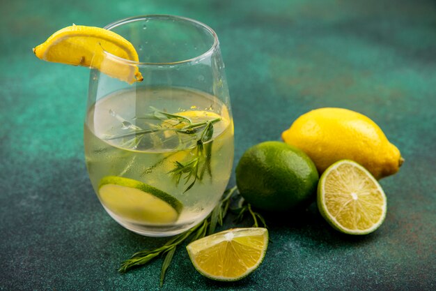 Вид сбоку освежающей воды для детоксикации в стакане на деревянной кухонной доске с ломтиками лайма, лимонов и палочками корицы на зеленом