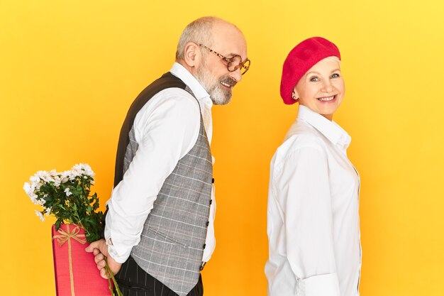 Side view of romantic cute senior man in elegant clothes holding flowers and box of gift behind his back making surprise present to stylish female who suspects nothing. Romance and Valentine's Day