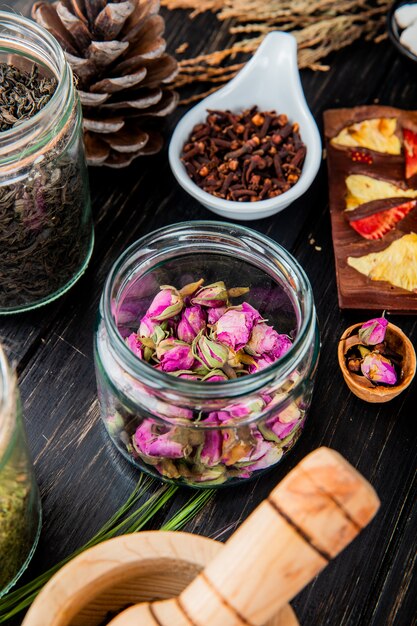 Side view of rose buds in a glass jar, dry black tea leaves, clove spice and chocolate bar with fruits on black wood