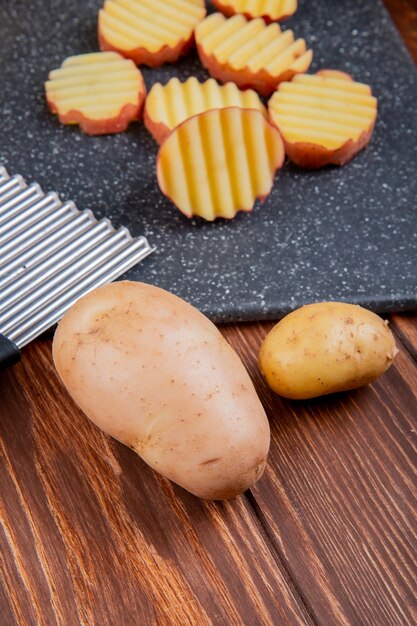 Side view of ruffled slices of potato on cutting board with whole ones on wooden table