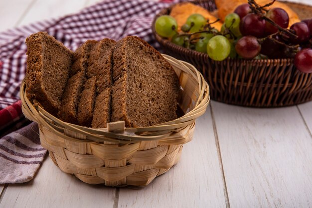 Side view of rye bread slices in basket on plaid cloth with basket of grapes nectacots on wooden background