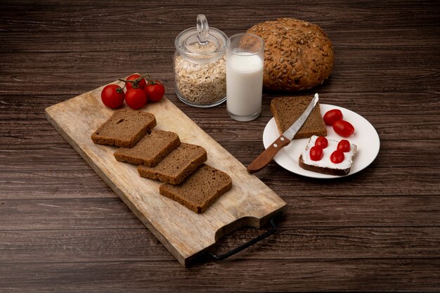 Side view of rye bread slices and tomatoes on cutting board with milk cob and oat-flakes on wooden background