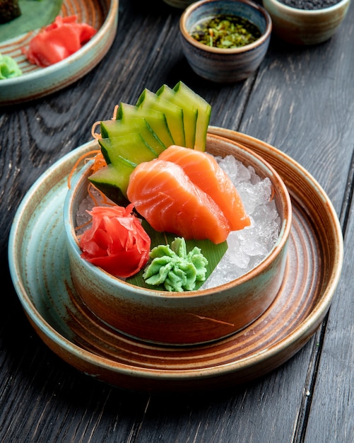 side view of salmon sashimi with sliced cucumbers ginger and wasabi sauce on ice cubes in a bowl on wooden table