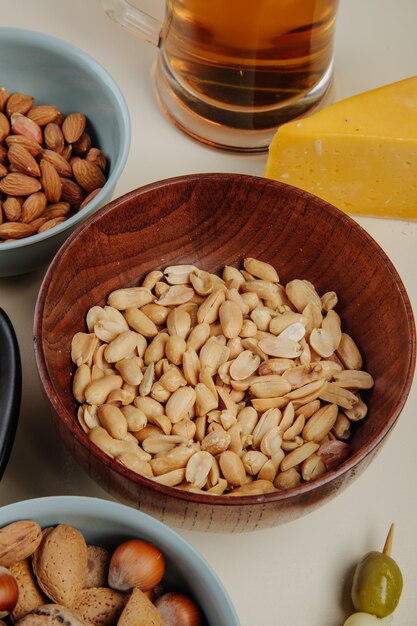 Side view of salted peanuts in a wood bowl with a piece of cheese and a glass of beer on white