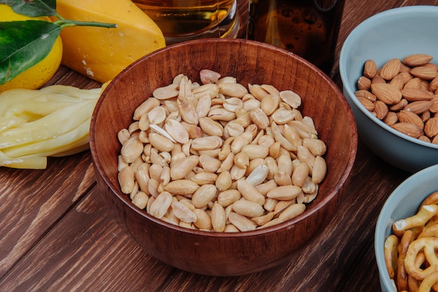 Vista laterale delle arachidi croccanti dello spuntino salato in una ciotola di legno e formaggio con birra su rustico