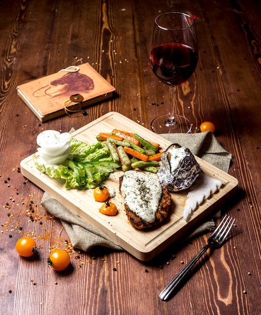 Free photo side view schnitzel with cream cheese with baked potatoes in foil and with vegetables on a board and a glass of red wine