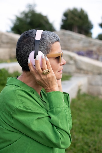 Side view of senior woman listening to music in park. Female model with short grey hair in bright clothes and big headphones. Leisure, activity, technology concept