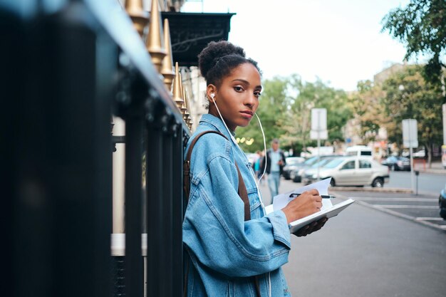 Side view of serious casual African American student girl in denim jacket with notepad confidently looking in camera on city street