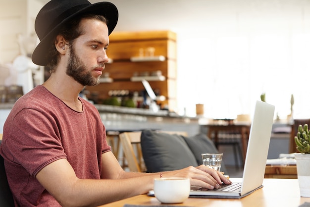 Side view of serious and concentrated bearded freelancer in black headwear keyboarding on laptop pc, working remotely, using free high-speed internet connection during breakfast at modern cafe