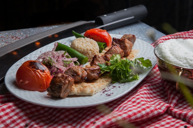 Вид сбоку шашлык с жареными овощами и нарезанным луком и айраном и ножом в белой тарелке