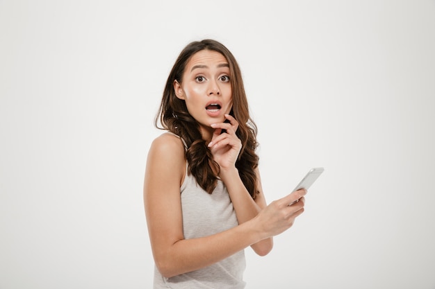 Side view of Shocked brunette woman holding smartphone and looking at the camera over gray