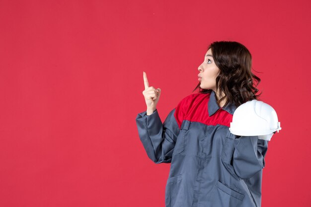 Side view of shocked female architect holding hard hat and pointing up on isolated red background