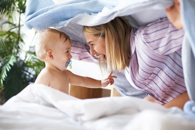 Side view shot of charming joyful young woman in pajamas playing hide and seek with toddler daughter. Adorable cute infant child sucking on pacifier looking at mother, having playful facial expression
